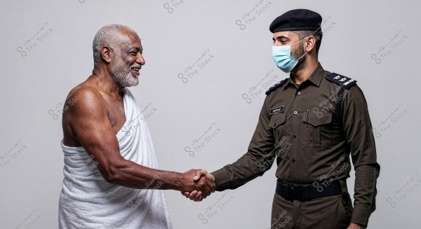 An image of two men shaking hands. The man on the left is wearing a white ihram, and the man on the right is dressed in a brown military uniform with a cap, wearing a blue face mask. The setting appears to be of Saudi context.
