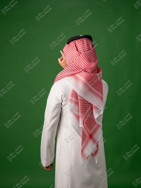 A man wearing a Saudi thobe and ghutra stands in front of a green background, with his face not visible.