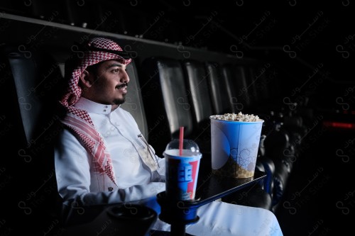 شاب سعودي يرتدي الزي السعودي التقليدي يجلس في صالة السينما ويظهر أمامه الفشار وأحد المشروبات على الطاولة