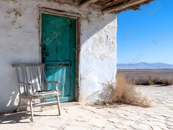 An image depicting a light blue wooden door set in an old white wall. Next to the door, there is an old wooden chair positioned on an uneven stone floor. In the background, a desert landscape with distant mountains under a clear blue sky is visible. Some dry shrubs are next to the wall.