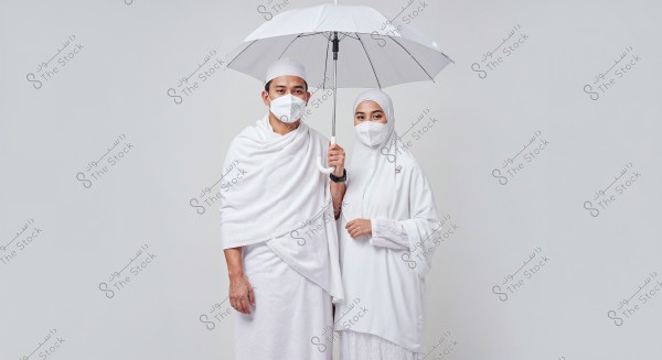 Image of a man and woman wearing traditional white Ihram attire and holding a white umbrella. Both are wearing face masks. The woman is wearing a white headscarf. The background is plain white, emphasizing the couple.