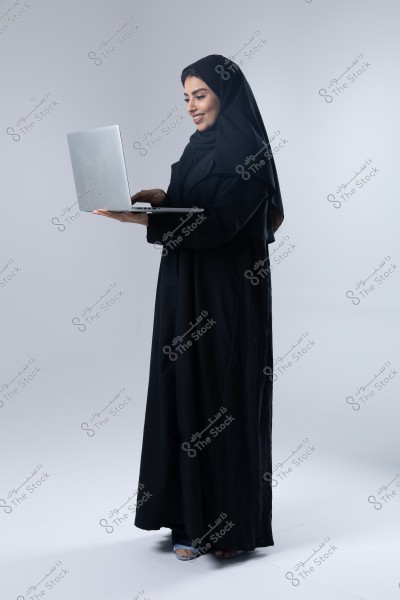 An image of a woman wearing a black abaya and hijab, standing and smiling while holding a silver laptop. The background is white, highlighting the woman. The attire suggests that the image might be from an Arab country, possibly Saudi Arabia.