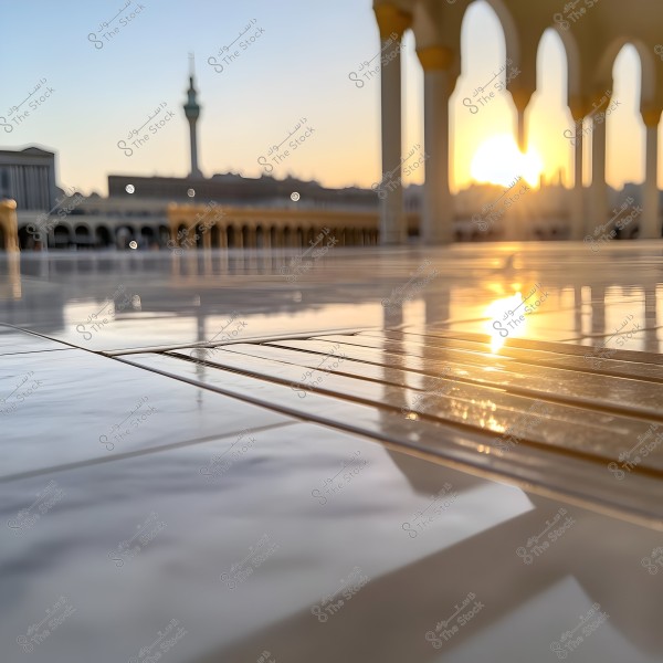 لقطة لأرضية رخامية في باحة مسجد كبير عند شروق الشمس, حيث يظهر انعكاس ضوء الشمس على السطح اللامع. في الخلفية, تظهر بعض الأقواس المعمارية وعِمارة المسجد والمئذنة تحت السماء الصافية.