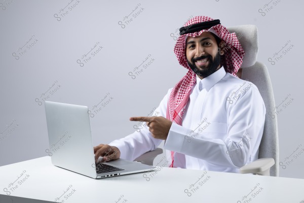 The image shows a man sitting on a comfortable chair behind a white desk, pointing his finger at an open laptop screen in front of him. The man is wearing traditional Saudi attire, including a white thobe, a red and white shemagh, and a black agal.