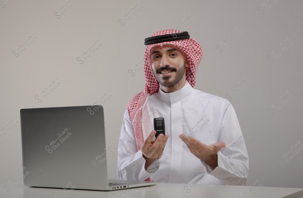 A man wearing a traditional Saudi thobe and red shemagh sits in front of a laptop. He holds a small remote control in his hand and appears to be smiling. The background is neutral with a light gray color.
