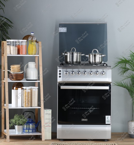 Image of a modern kitchen featuring a silver \"Bison\" oven with a stovetop, with several metallic pots placed on it. Next to it is a wooden shelf with multiple tiers displaying various kitchen supplies like grains, pasta, and oil in bottles and containers. A part of a plant is visible on the right, adding a touch of nature to the scene. The floor is partially visible around the oven.