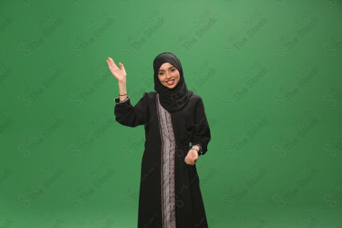 Saudi woman on green background wearing abaya and hijab, making gestures with her hands, raising her hand smiling, Eid celebrations