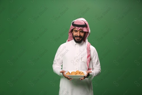 شاب سعودي على خلفية خضراء يرتدي الثوب و الشماغ، يقوم بعمل حركات بيده،يمسك بصينية الكعك و يقدمه، احتفالات العيد