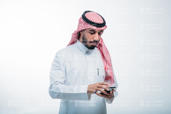 رجل سعودي يرتدي الزي الرسمي السعودي على خلفية بيضاء ، يمسك الجوال ، يستخدم أحد تطبيقات الآيفون ، يتصفح الإنترنت ، يستخدم وسائل التواصل الاجتماعي