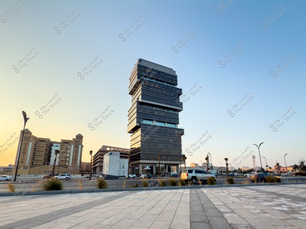 منظر لمبنى حديث ذو تصميم معماري مميز يتكون من عدة طوابق مكدسة بشكل غير تقليدي, يقع بجانب طريق عام مع وجود سيارات متوقفة ومتحركة. في الخلفية يظهر مبنى فنادق عليه اسم "وارويك". السماء صافية مع ضوء شمس الغروب الذي يضفي وهجاً دافئاً على المشهد.