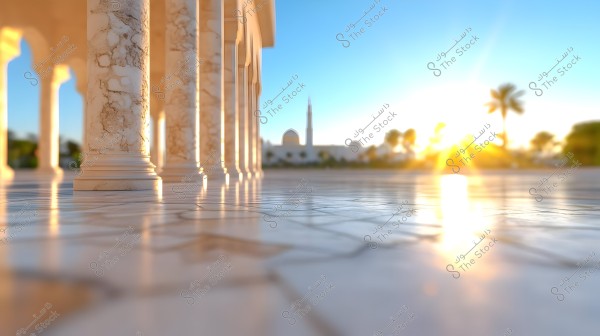 The image depicts an architectural scene featuring detailed marble columns with prominent carvings, along with a shiny floor reflecting the bright sunlight. In the background, a mosque with its dome and palm trees in an open area can be seen, with the horizon illuminated by the setting sun\'s light.