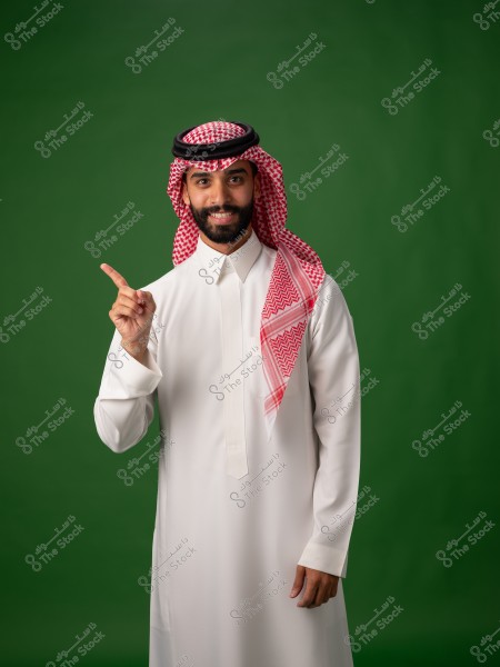 A portrait of a man wearing traditional Saudi attire, featuring a white thobe, a red and white shemagh, and a black agal. He stands against a green background, smiling and pointing upwards with his index finger.