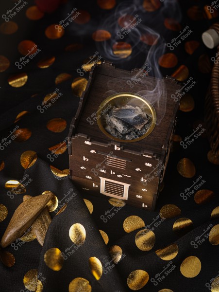 Image of incense burning inside a traditional wooden incense burner with smoke rising from it. The burner is placed on a black fabric decorated with shiny golden dots. Next to the burner, there are two pieces of aromatic wood.