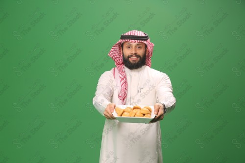 شاب سعودي على خلفية خضراء يرتدي الثوب و الشماغ، يقوم بعمل حركات بيده،يمسك بصينية الكعك و يقدمه، احتفالات العيد