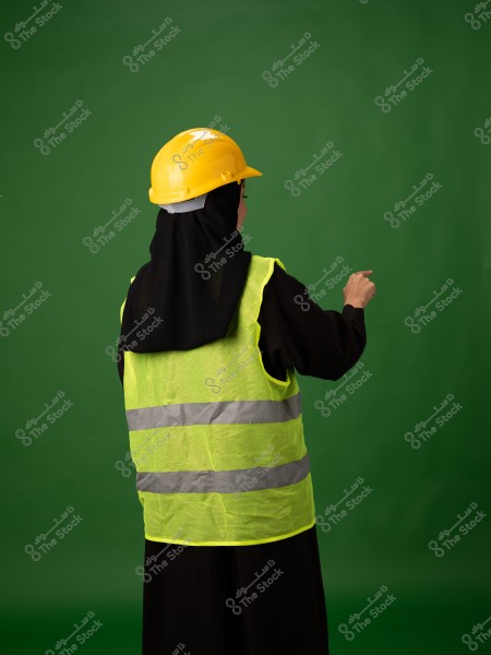 An image of a woman wearing a yellow reflective vest and a yellow safety helmet. The woman is shown from the back, also wearing a black hijab. The background is green, creating a striking contrast with the colors. She appears to be working in construction or safety.
