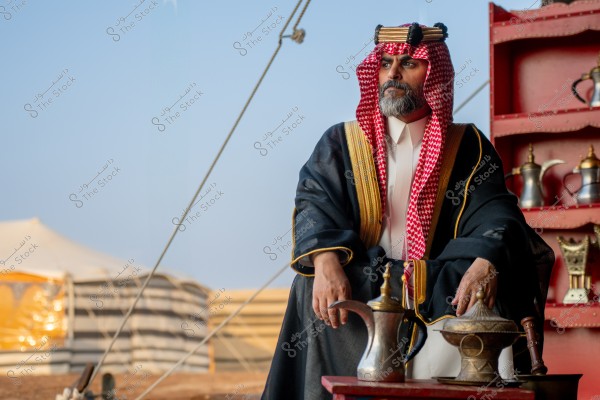 A man wearing traditional Saudi attire is sitting beside a tent. He is dressed in a ghutra and agal, along with a traditional thobe, and sits next to a table with Arabic coffee pots. In the background, a tent is set up in the desert.
