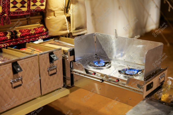 The image shows a metal double burner stove with a blue flame, placed on a makeshift kitchen surface. Next to it are brown padded storage boxes, and in the background, there are colorful and embroidered fabrics with traditional designs. The scene suggests a camping or outdoor cooking environment.
