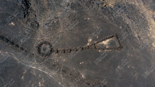 An aerial image of an archaeological site in a desert region, featuring a large stone formation shaped like a circle connected to a rectangle by a straight line. The formation appears ancient and is situated on barren, rocky terrain.