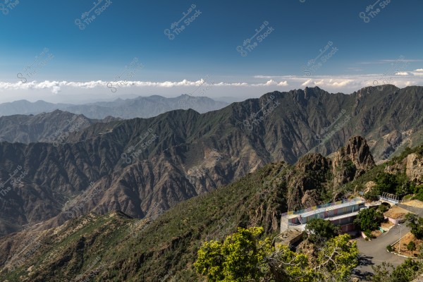 A stunning natural landscape of lofty mountains under a clear blue sky. The mountains, covered in green vegetation, stretch across the horizon with some distant white clouds in the sky. In the foreground, there is a small multicolored building showcasing architectural details situated near the edge of the rocks.