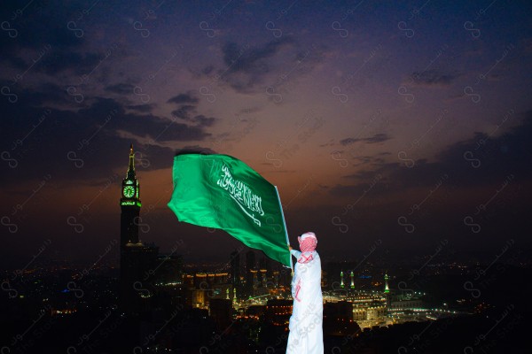 A snapshot of a Saudi man wearing the traditional Saudi dress, carrying the Saudi flag over a mountain in the Makkah Al-Mukarramah region, and the sky appears clear, the royal clock tower building in the Grand Mosque, buildings and landmarks, the Grand Mosque of Mecca.