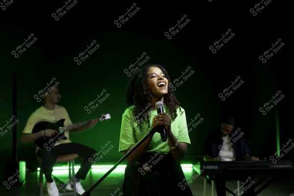 امرأة تغني في استوديو بينما يعزف رجلان على الجيتار والبيانو في الخلفية.