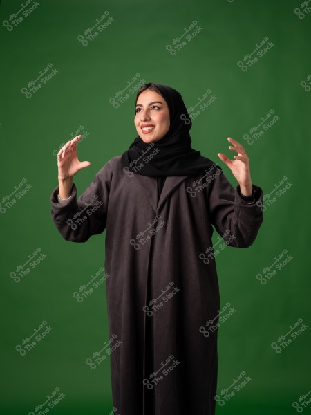 A portrait of a woman standing against a green background. She is wearing a black abaya and hijab. Her hands are raised in a gesture as if she is expressing or pointing at something. Her facial expression shows a smile and optimism.