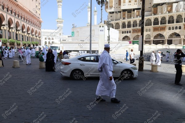 The image shows an outdoor scene in the area of the Holy Mosque in Mecca, Saudi Arabia. Many people are wearing traditional white garments and black abayas, indicating their participation in Umrah or Hajj rituals. In the background, Islamic architectural buildings and minarets are visible. A white car is parked in the center, with people walking around it.