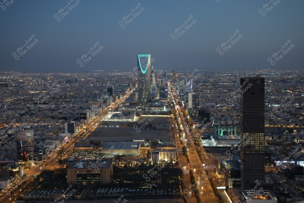 A night view of Riyadh, Saudi Arabia, showcasing the Kingdom Tower illuminated with distinctive blue lights in the city center. The streets are lit by car lights, and the surrounding buildings create a vibrant urban scene.
