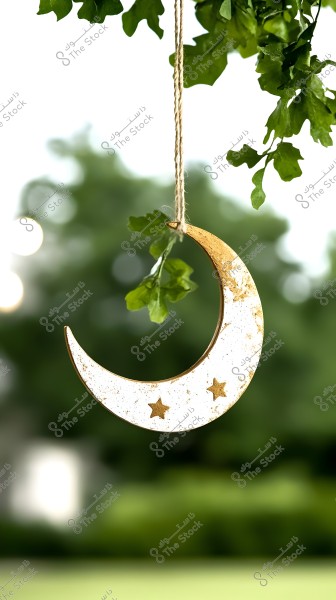 Image depicts a golden crescent moon hanging by a string from a tree, featuring two small stars. The background shows a blur of green leaves, imparting a sense of nature and tranquility.