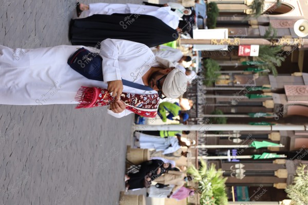 The image shows a man wearing a white thobe and traditional head covering, holding a decorated piece of cloth. He is wearing a black cross-body bag. In the background, there are people dressed in traditional clothing and green flags indicating Saudi Arabia, along with modern buildings and palm trees.