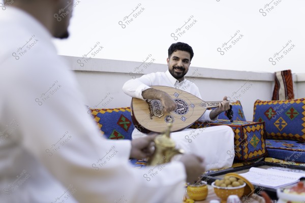 رجل يعزف على العود في جلسة تقليدية مع شخص يحمل دلة قهوة عربية, مع وسائد ملونة في الخلفية.