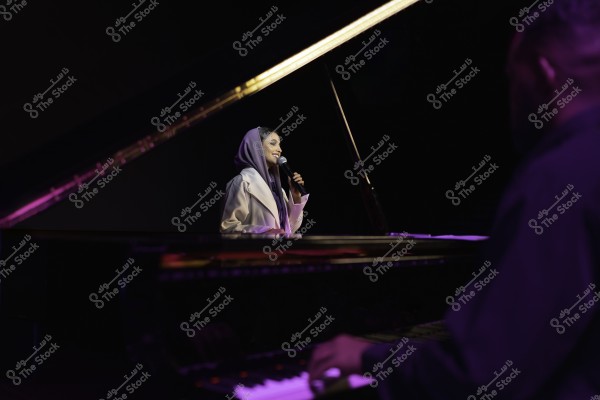 امرأة تغني على المسرح ممسكة بالميكروفون, بينما يعزف شخص آخر على البيانو في المقدمة.