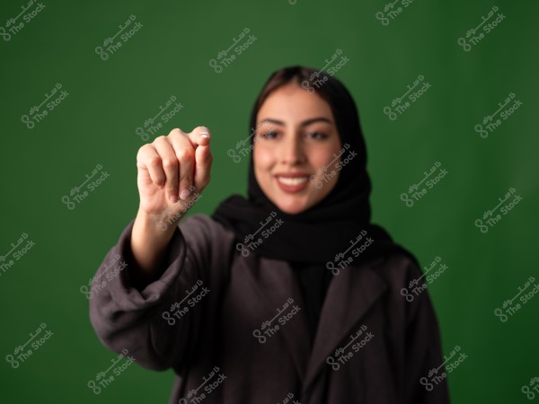 A portrait of a woman wearing a black hijab and a black coat against a green background. She is raising her hand and pointing with her finger while smiling.