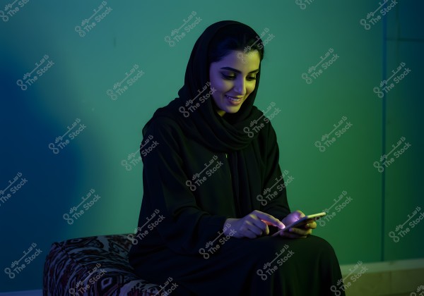 A portrait of a woman wearing a black abaya and hijab, looking at her smartphone with a smile in a room lit with blue and green hues. She is seated on a traditionally patterned sofa.