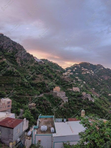 A scenic view of a village situated on the side of a mountain covered with lush greenery. The houses are scattered along the slopes, with some partially constructed. The sky appears clouded with shades of orange and purple, indicating a sunset or sunrise. Buildings have roofs of various colors, including white and red, and green trees are spread throughout the area.