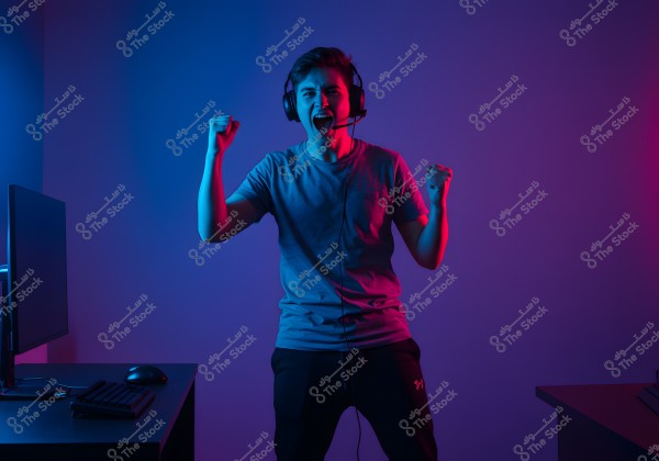 Image of a young man wearing a headset standing in a room lit with blue and red colors, with his arms raised excitedly. He is wearing a gray t-shirt and dark sport pants. In the foreground, there is a desk with a computer and a keyboard.
