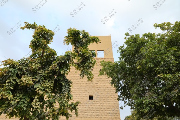 Image showing a tower constructed from brick with a yellow facade, surrounded by lush green trees. The tower features square windows and is set against a cloudy sky.
