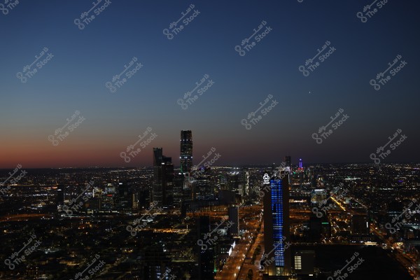 Image of the skyline of Riyadh, Saudi Arabia, at dusk. The sky is a blend of blue and faint orange colors. Tall skyscrapers are illuminated, notably the Kingdom Tower and other glowing buildings, with streets filled with cars and light.
