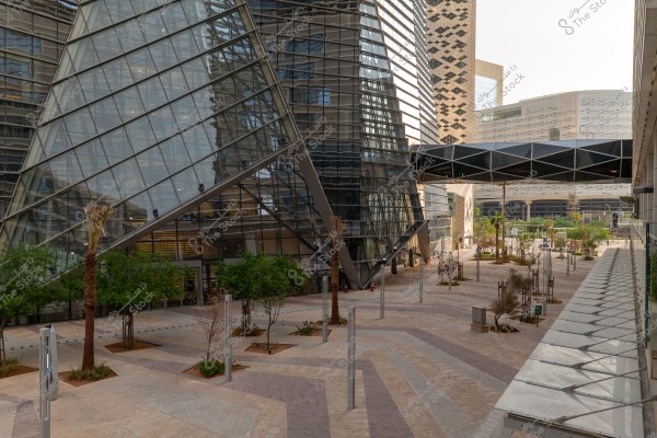 The image depicts an urban scene featuring a modern building with a large glass structure in the foreground. There is an overhead walkway connecting the buildings. Some trees and plants are visible in the open space with lamp posts. The background includes other buildings with contemporary designs under a clear sky.
