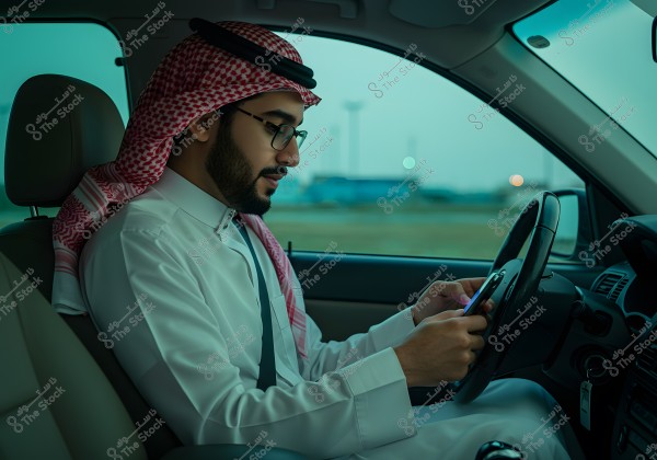 Image of a man sitting in the driver\'s seat of a car, wearing traditional Saudi clothing including a thobe, a ghutra, and an agal. The man is looking at a mobile phone held in his hands. The scene captures an interior car setting with a natural light background outside.