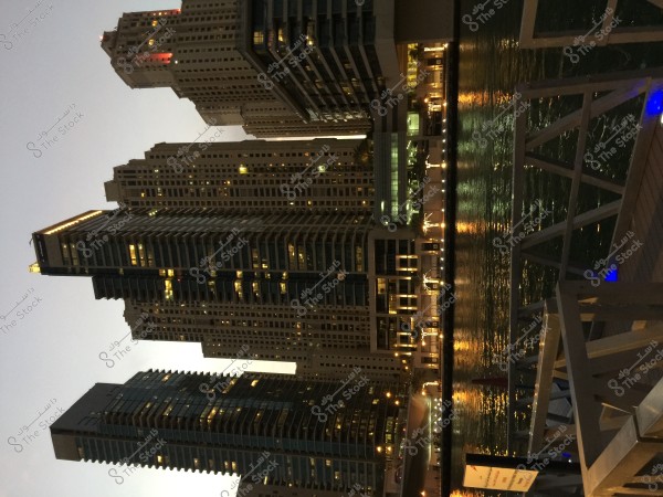 A nighttime image of a group of skyscrapers in an urban city, with illuminated interior lights creating a beautiful skyline. The nearby water reflects the lights, enhancing the beauty of the scene. Part of a metal structure is visible in the foreground beside the water.
