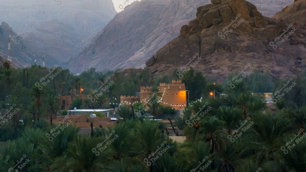 منظر طبيعي لقرية عقدة في حائل وسط واحة نخيل مع جبال مرتفعة في الخلفية. المباني ذات الطراز المعماري التقليدي مكونة من طين وأعمدة خشبية, وتحيط بها أشجار النخيل الكثيفة وتزينها أضواء صغيرة. يمتد الوادي بين الجبال في الخلف, ما يضيف عمقاً للمشهد.