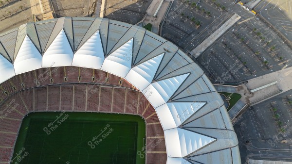 An aerial view of a large sports stadium with a modern architectural design, featuring a section of circular red seating and a white roof with consecutive triangular shapes. The stadium has a green field with surrounding parking spaces, some trees, and organized markings.