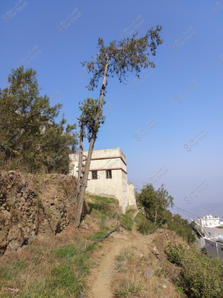 The image depicts a natural landscape featuring a two-story building with aged white walls situated on the edge of a hill. In the foreground, a narrow dirt path runs between rocks and wild vegetation. A tall, slender tree stands near the building. The horizon shows a wide expanse of blue sky with some distant mountains visible in the background.