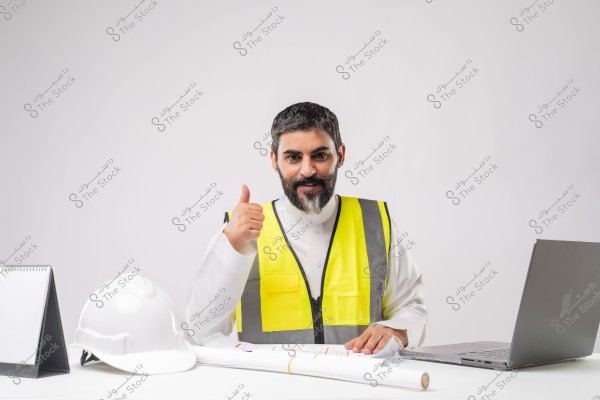 A portrait of a man sitting at a desk wearing a white thobe and a yellow safety vest. He is smiling and giving a thumbs-up, indicating approval. On the desk in front of him are a white helmet, blueprints, and a laptop. The traditional Arabic attire and safety vest suggest he works in construction or engineering in a region like Saudi Arabia.