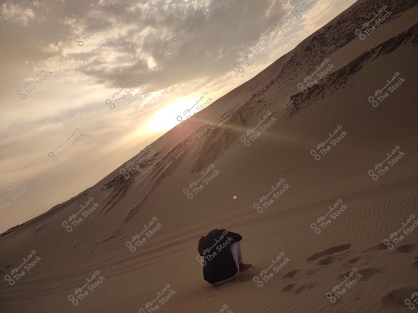 The image shows a wide desert landscape with sand dunes under a setting sun. A person is sitting on the sand in the foreground, looking towards the sunset. The sky is filled with thin clouds reflecting the sun\'s rays beautifully on the horizon.
