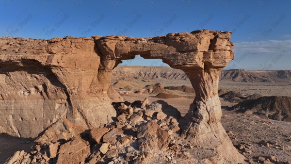 A natural landscape of a large rock formation shaped like an arch in a vast desert under a clear blue sky.