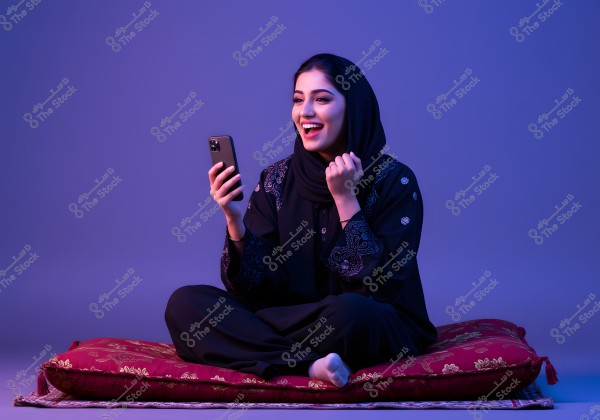 A portrait of a woman happily sitting on a traditional red patterned cushion, holding a smartphone. She is wearing a black abaya and hijab, with soft blue lighting in the background. The attire suggests the image may be from the Gulf region.