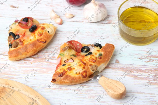 شريحتان من البيتزا موضوعة على سطح خشبي فاتح اللون, مُغطاة بالجبن والزيتون الأسود والطماطم وقطع اللحم. توجد أداة تقديم خشبية بجانب إحدى الشرائح. في الخلفية, يوجد فص ثوم وكوب زجاجي مملوء بزيت الزيتون.