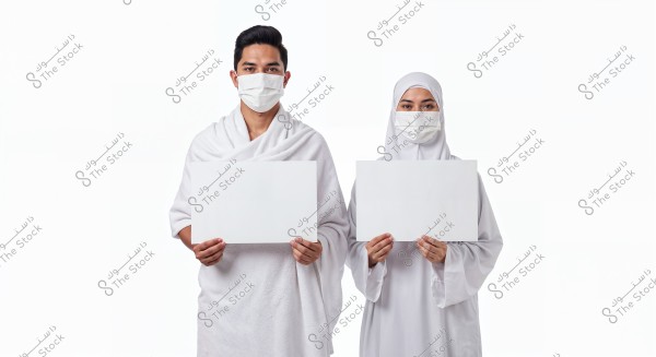 An image of a man and woman standing side by side, wearing traditional white Ihram clothing. Their faces are covered with medical masks, and each is holding a blank white sign. The background is white, enhancing the foreground details.